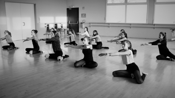 ten young women in a dance studio sit on the floor and reach their arms to the side